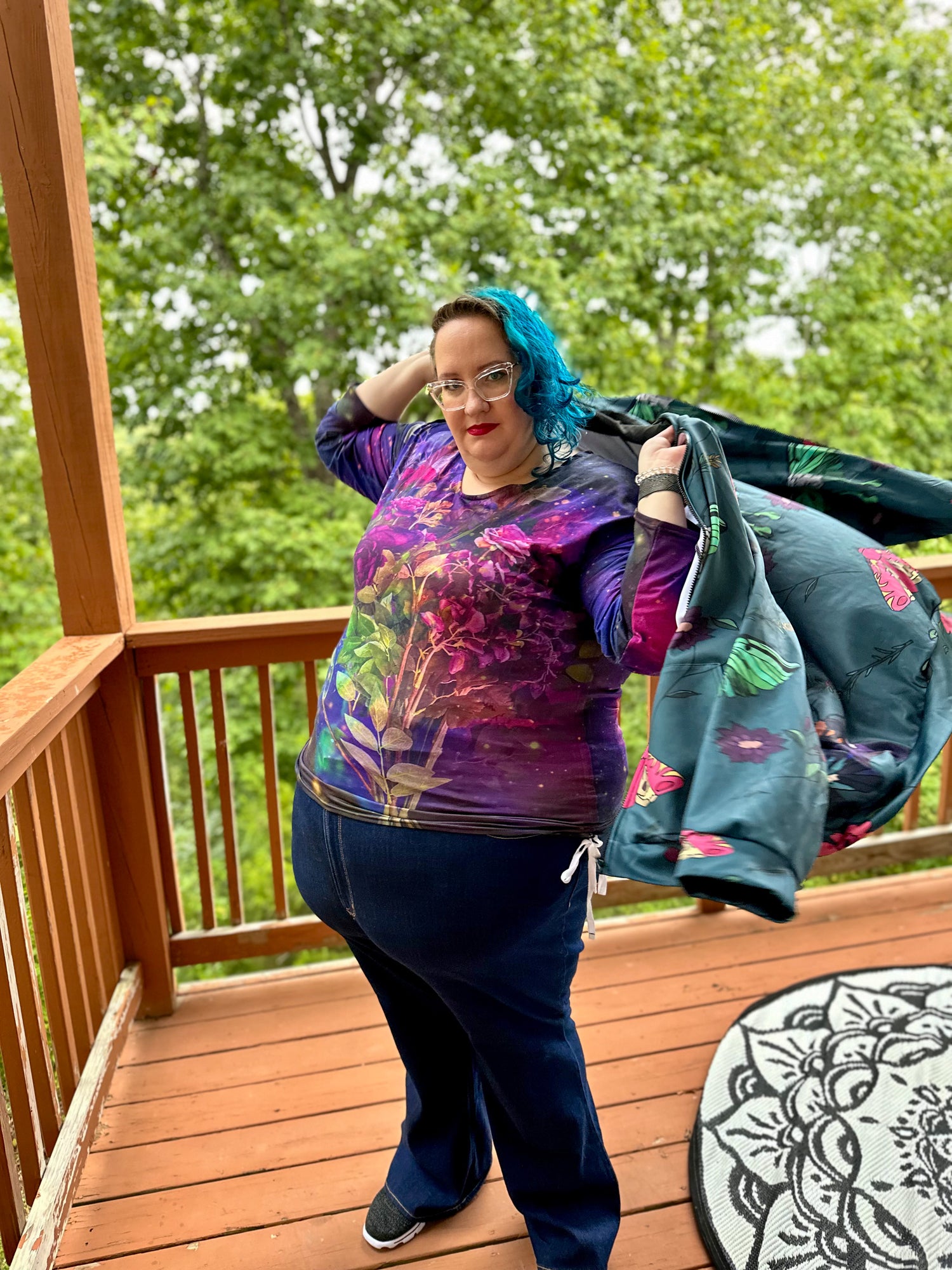 Woman standing on rust orange deck in jeans and a long sleeve shirt with flowers and galaxies. She is putting on a teal hoodie with flowers and winged things on it.